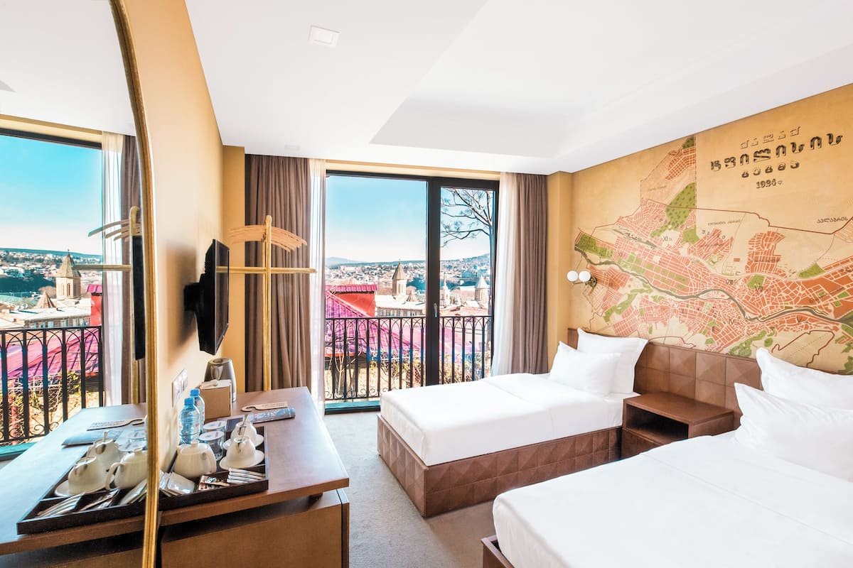 A hotel room with two beds, a desk, a chair, a map on the wall, and a balcony with a view of the city.