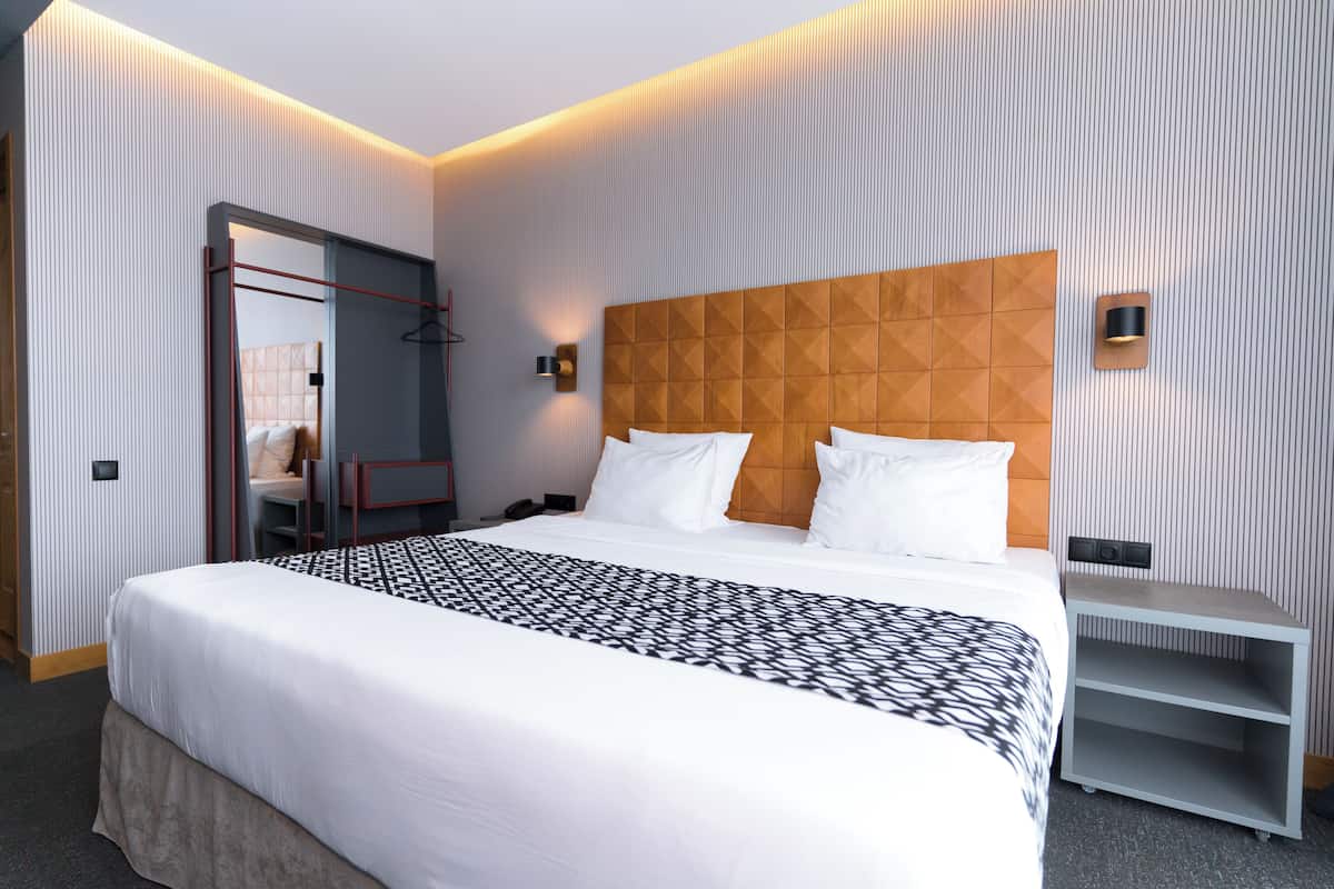 A modern hotel room with a large bed, a wooden headboard, a bedside table, and a wardrobe with a mirror.