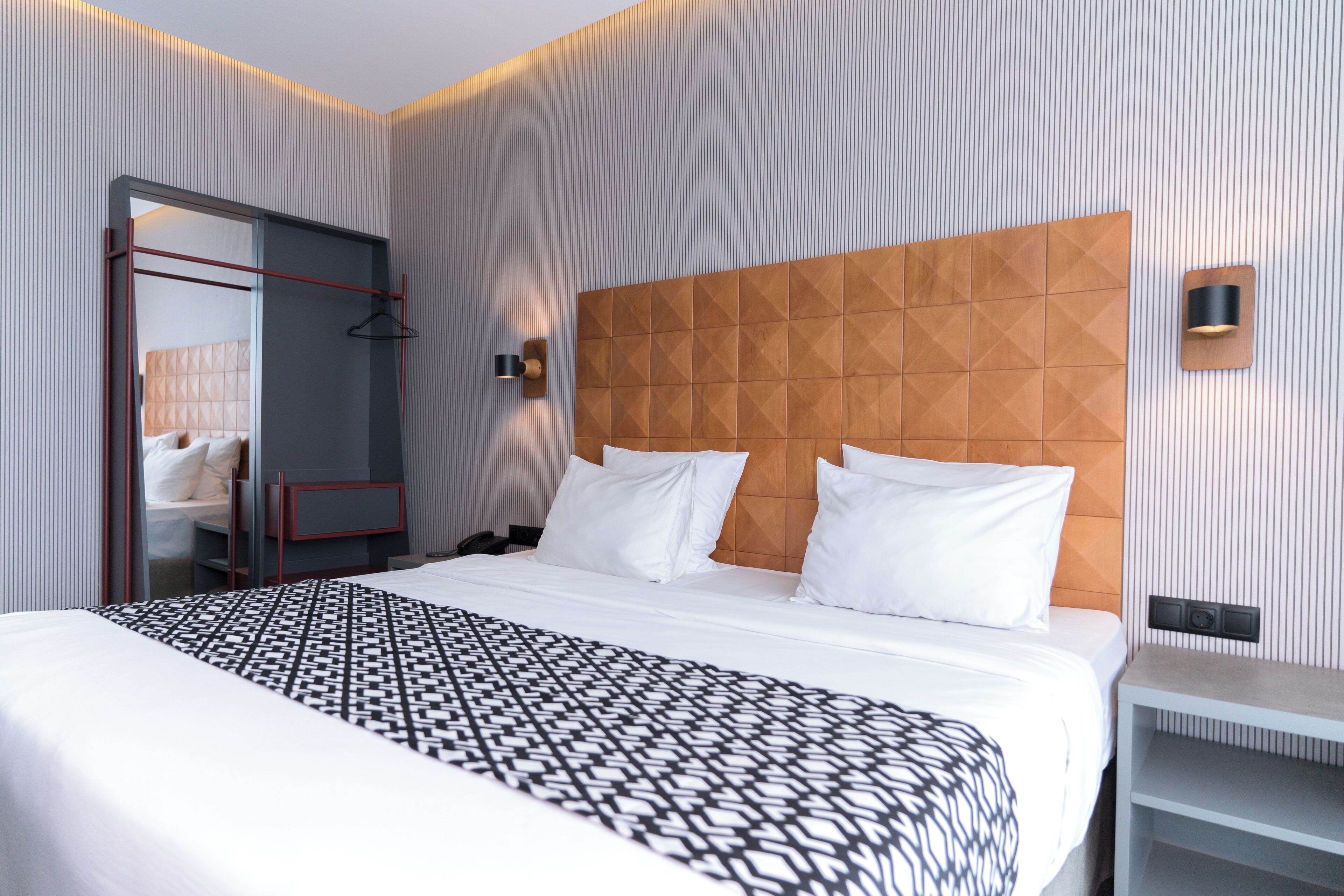 Une chambre d’hôtel moderne avec un grand lit, une tête de lit en bois, une table de chevet et une armoire avec un miroir.