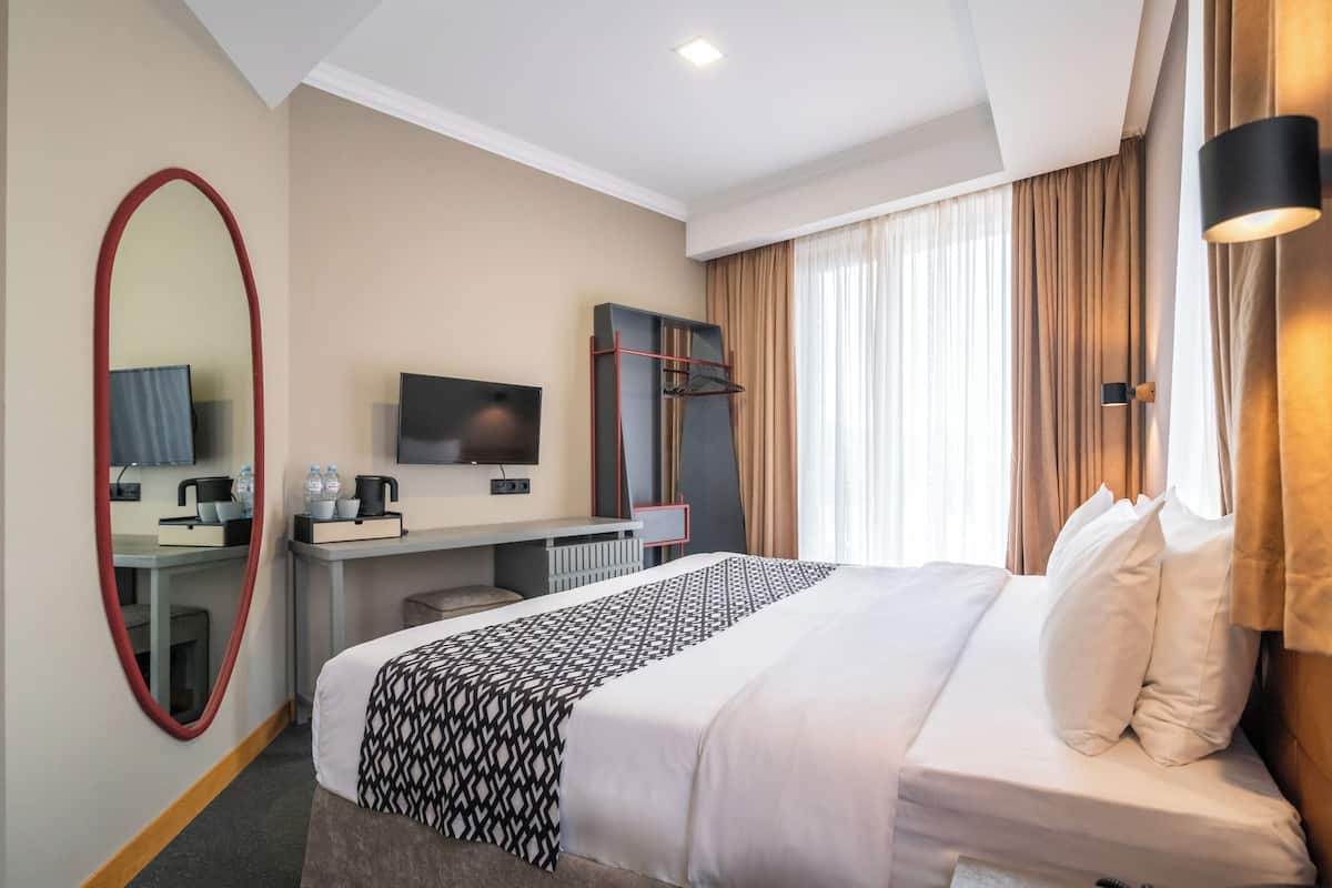 A hotel room with a large bed, a desk with a flat-screen TV, a mirror, and a window with curtains.
