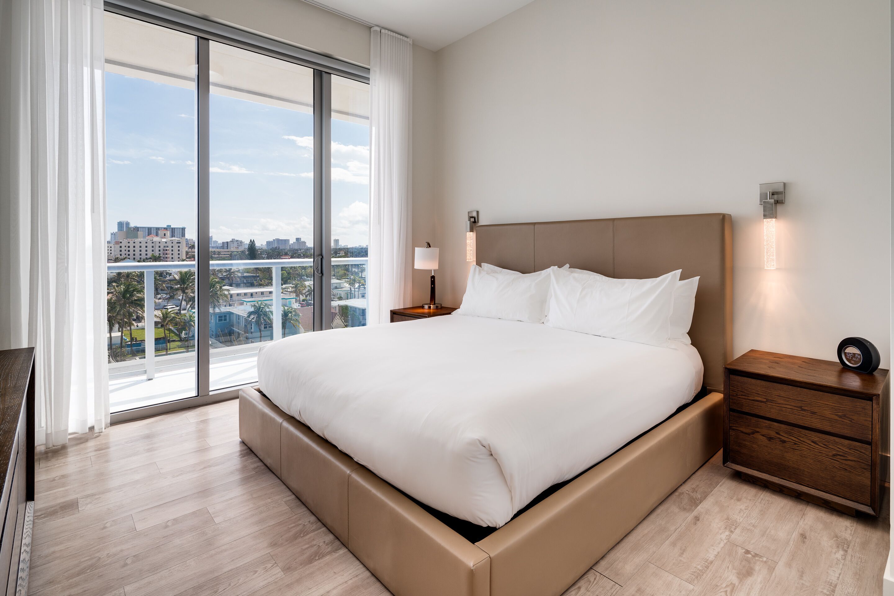 Moderni hotellihuone, jossa on suuri sänky, yöpöytä ja näkymä kaupunkiin lattiasta kattoon ulottuvien ikkunoiden kautta.