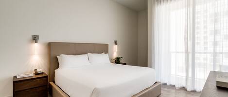 A modern bedroom with a large bed, bedside tables, and a view of the city through sheer curtains.