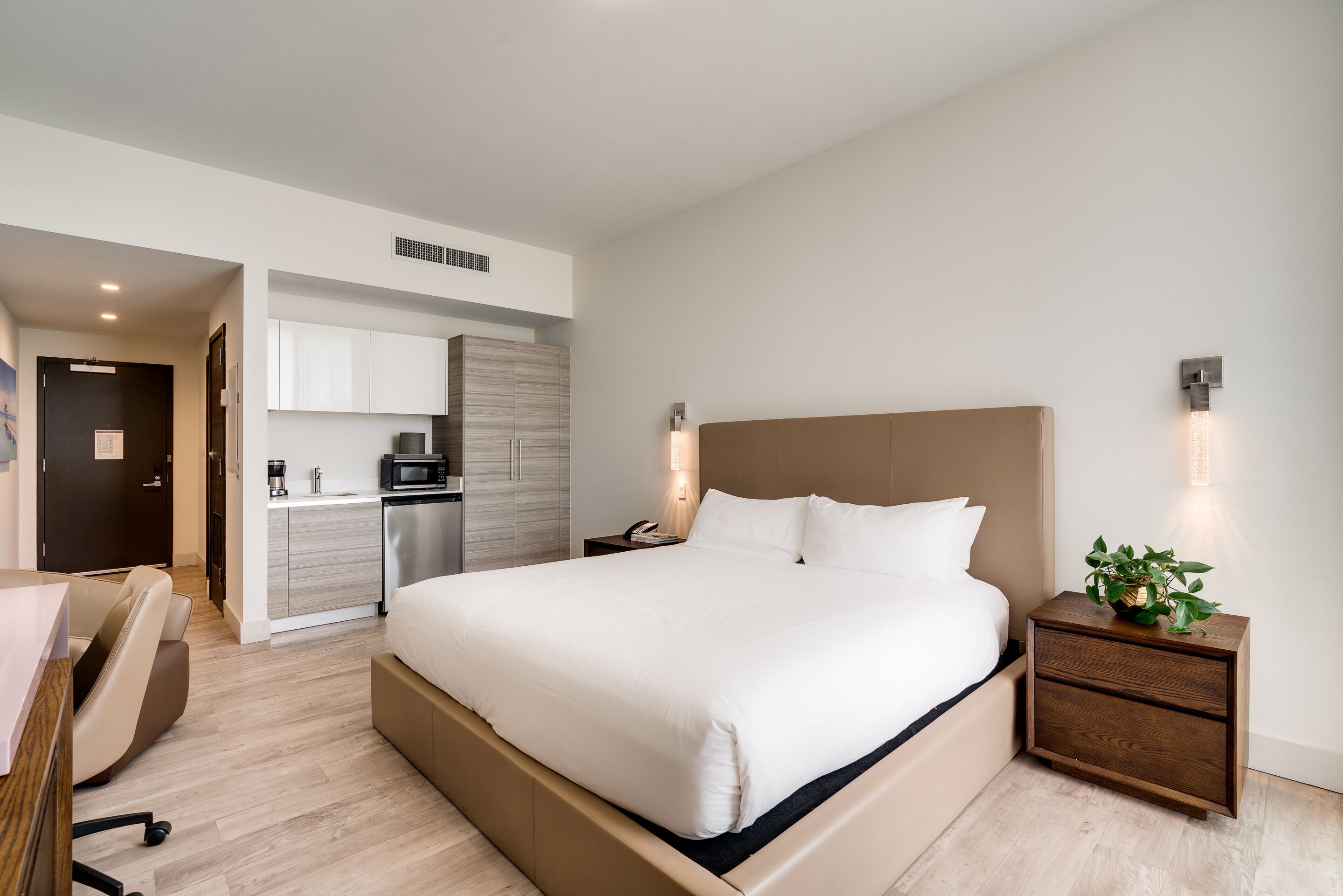 Camera d'albergo moderna con un letto grande, una scrivania, una sedia, un comodino con una pianta e una piccola cucina sullo sfondo.