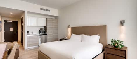 Camera d'albergo moderna con un letto grande, una scrivania, una sedia, un comodino con una pianta e una piccola cucina sullo sfondo.