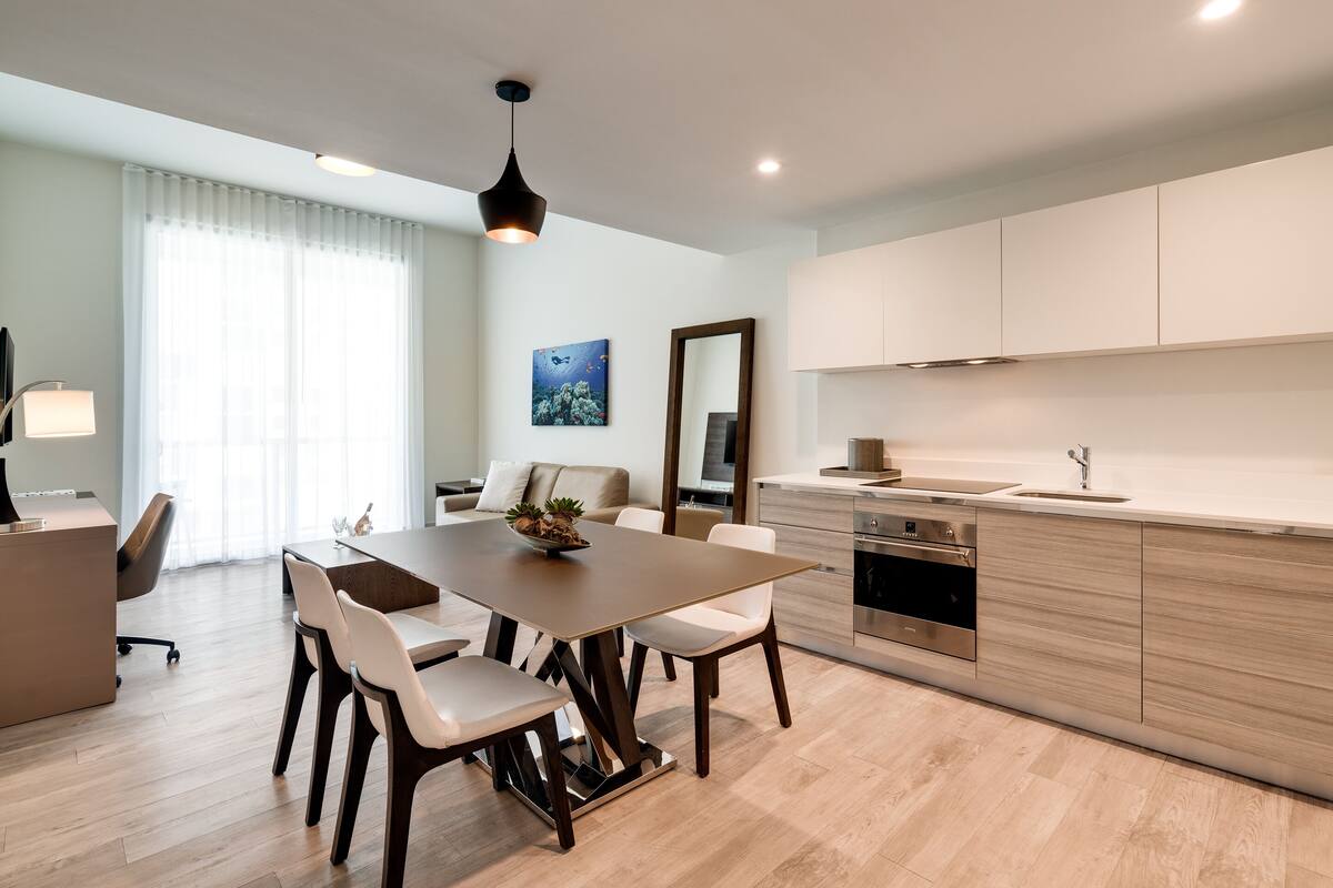Une cuisine moderne avec un coin repas, comprenant une table en bois, des chaises blanches et une grande fenêtre agrémentée de rideaux légers.