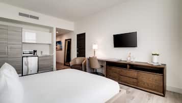 Quarto de hotel moderno com uma cama grande, cômoda de madeira, escrivaninha com luminária, televisão de tela plana e cozinha compacta com micro-ondas e cafeteira.