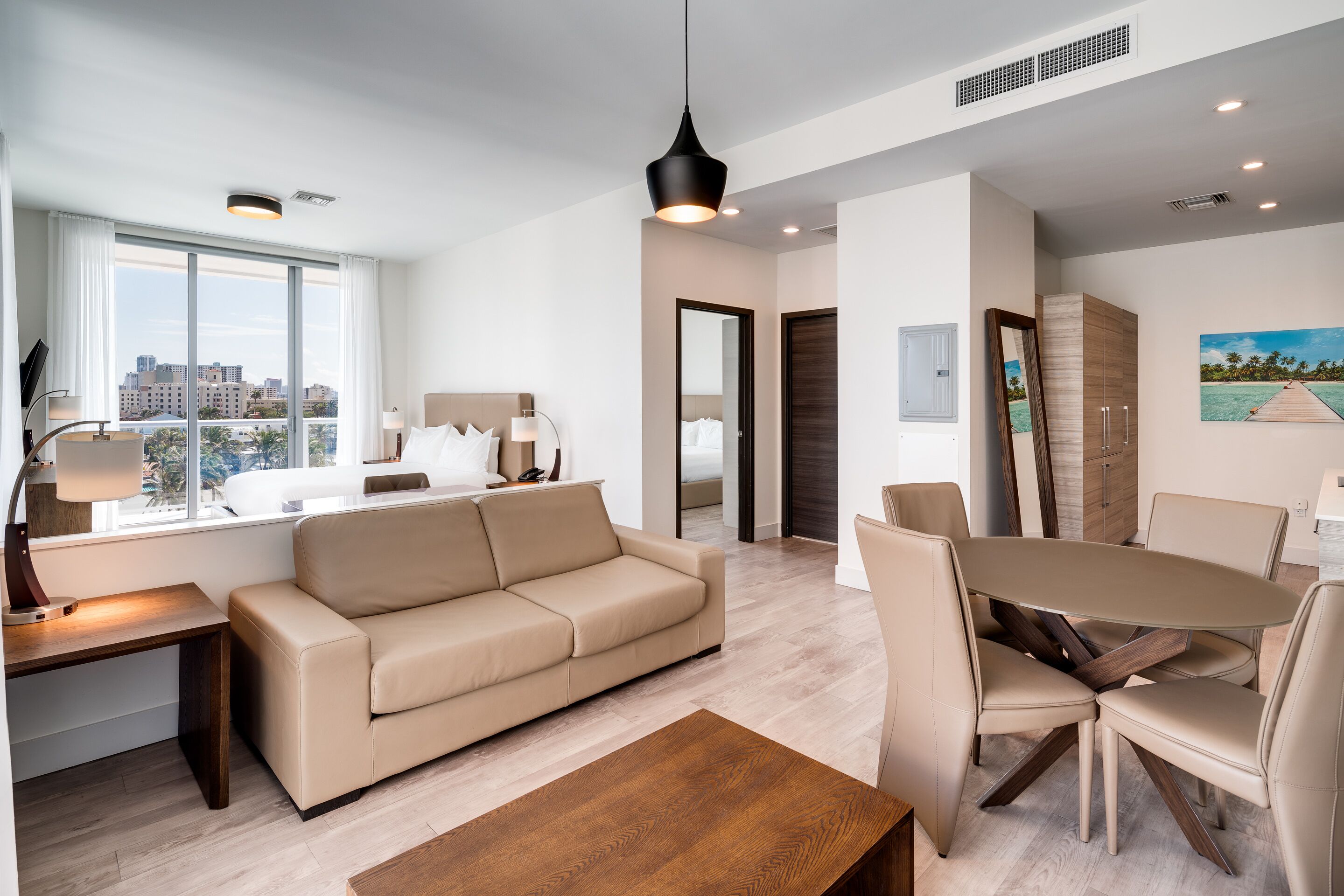 A modern living room with a sofa, coffee table, and dining area. There is a large window with a city view, a door leading to another room, and a painting on the wall.