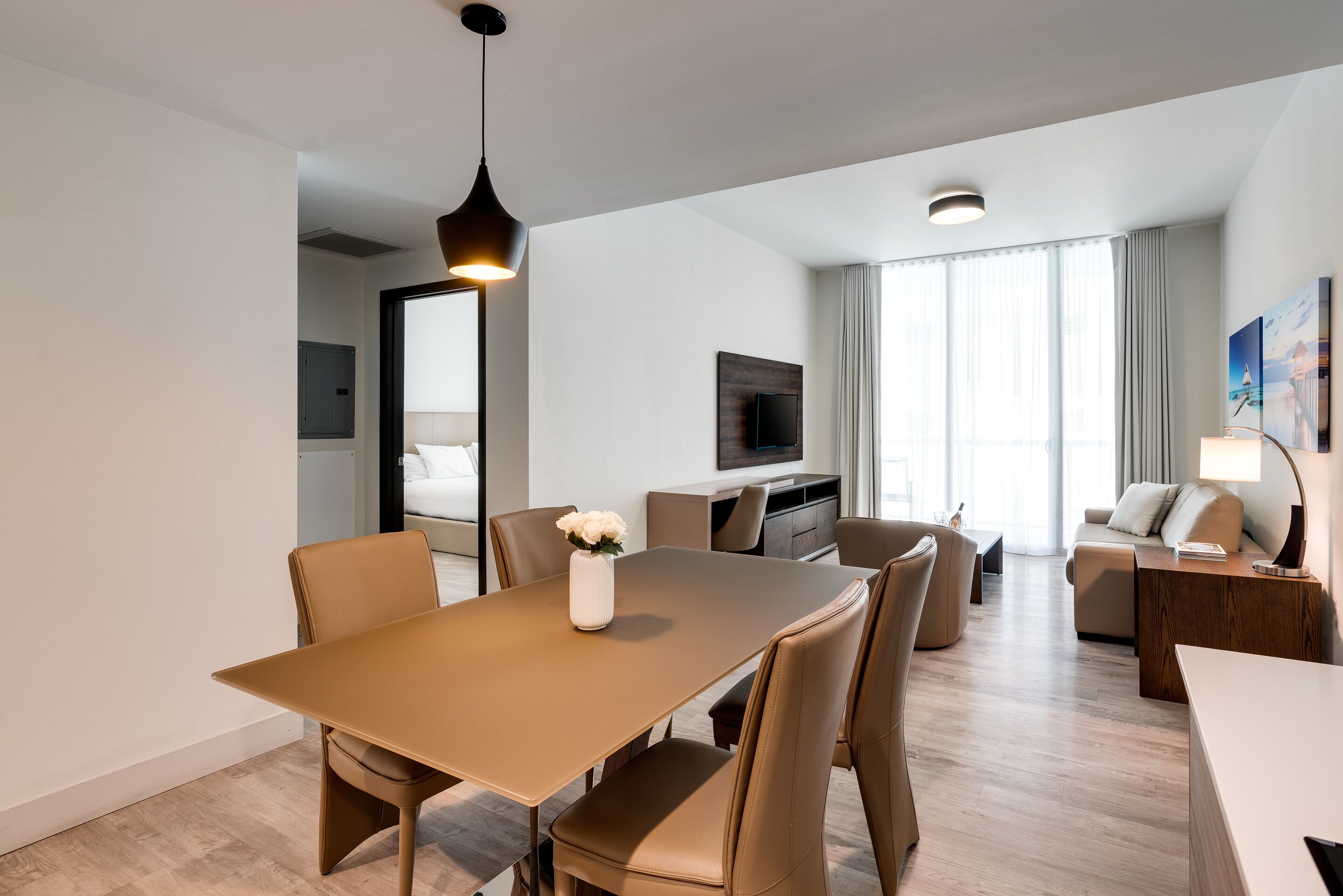 Et moderne hotelværelse med spiseplads, sofa, fjernsyn og et stort vindue med gardiner.