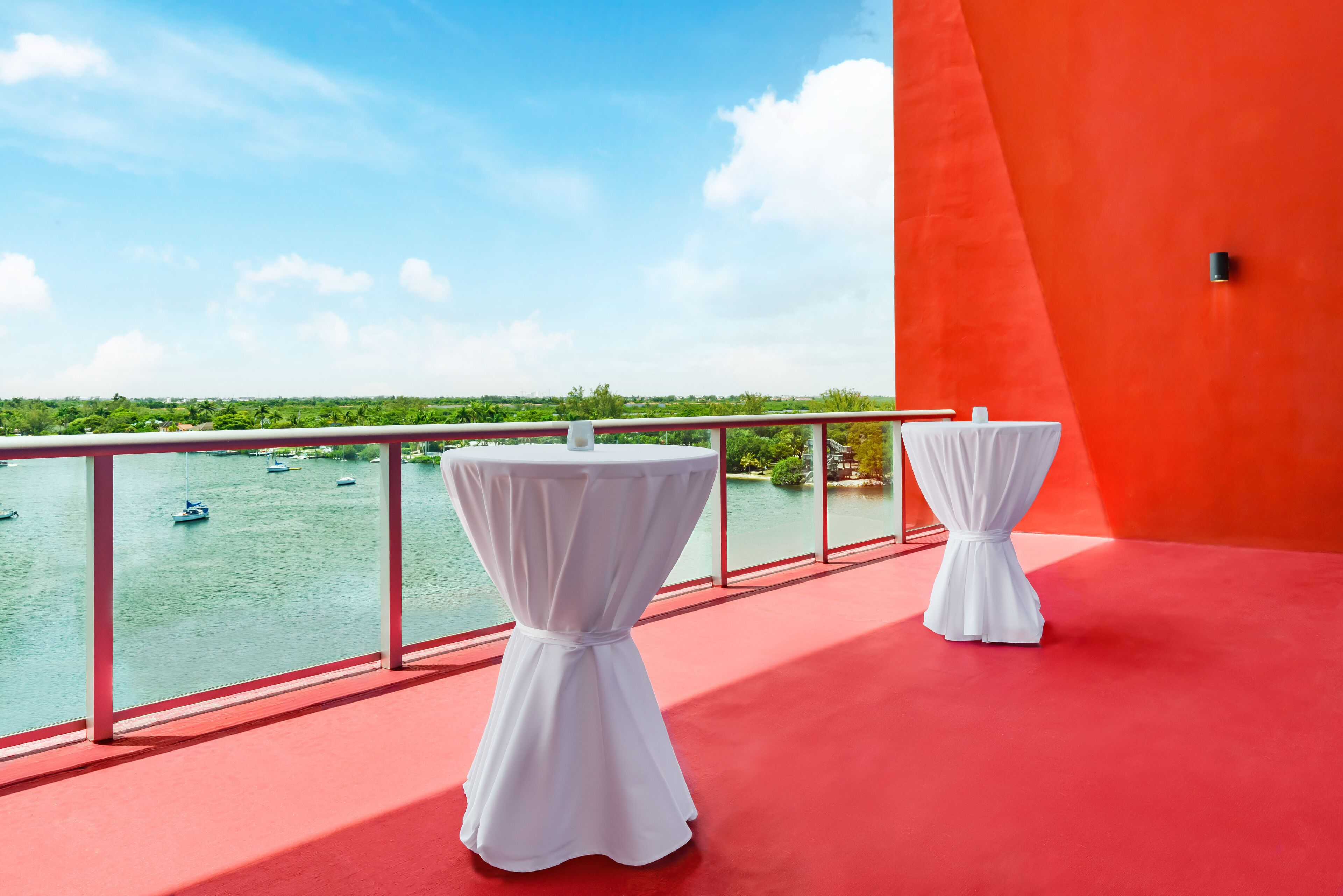 Un balcon avec deux tables blanches, un sol rouge et une vue sur des bateaux sur l’eau.