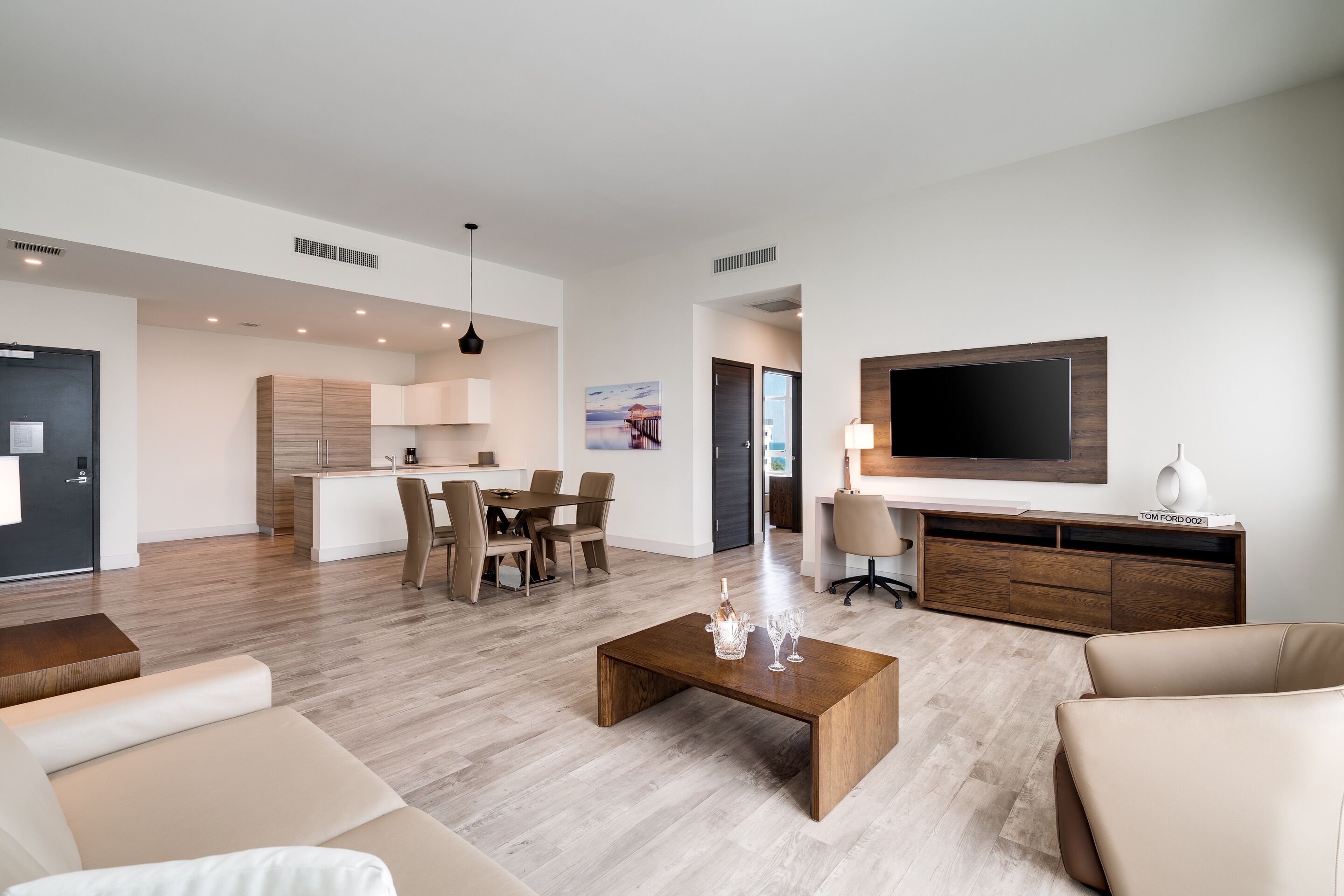 A modern living room with a large flat-screen TV, a coffee table, and a dining area with chairs and a kitchen in the background.