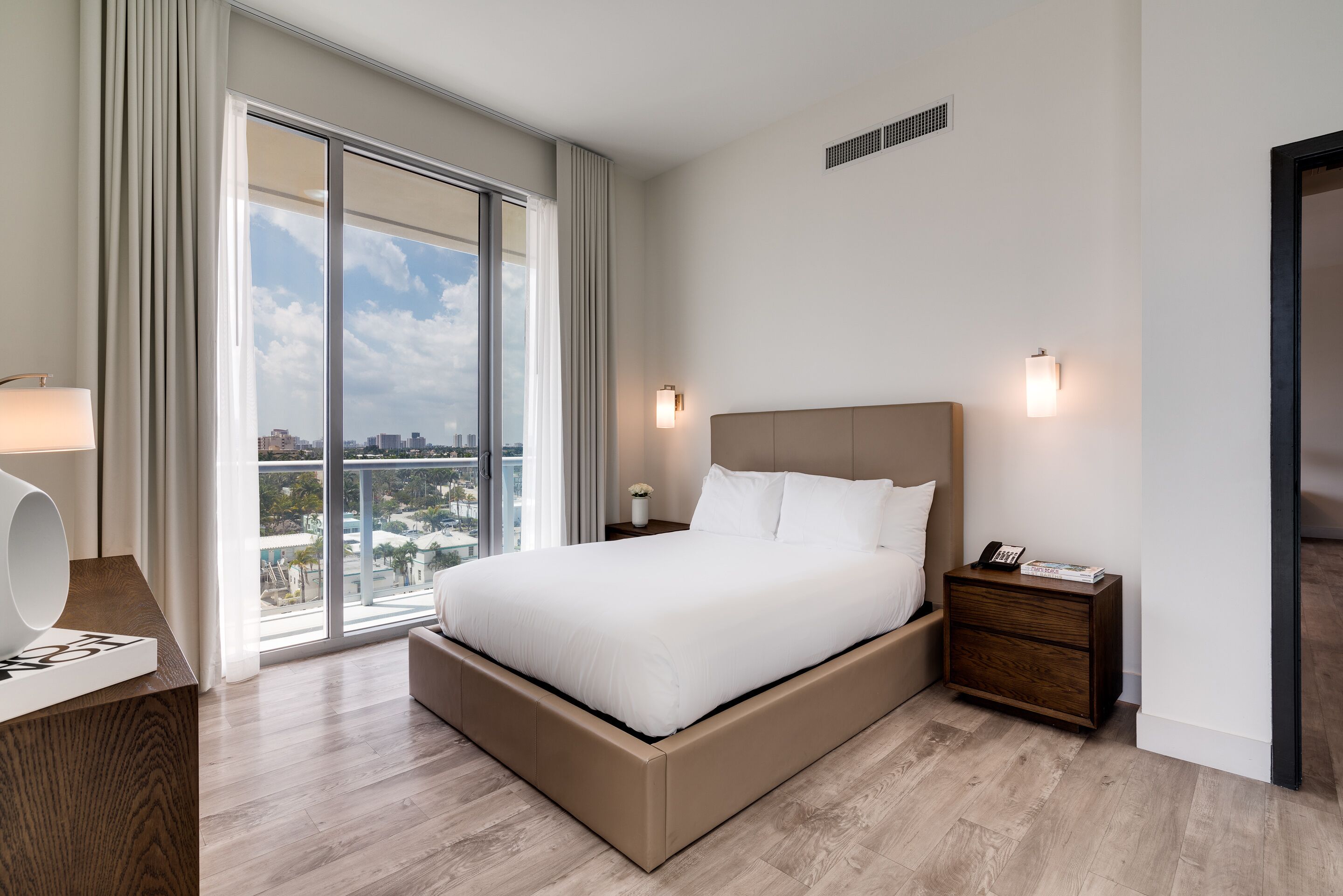 A modern hotel room with a large bed, a nightstand, a lamp, and a view of the city through large windows.