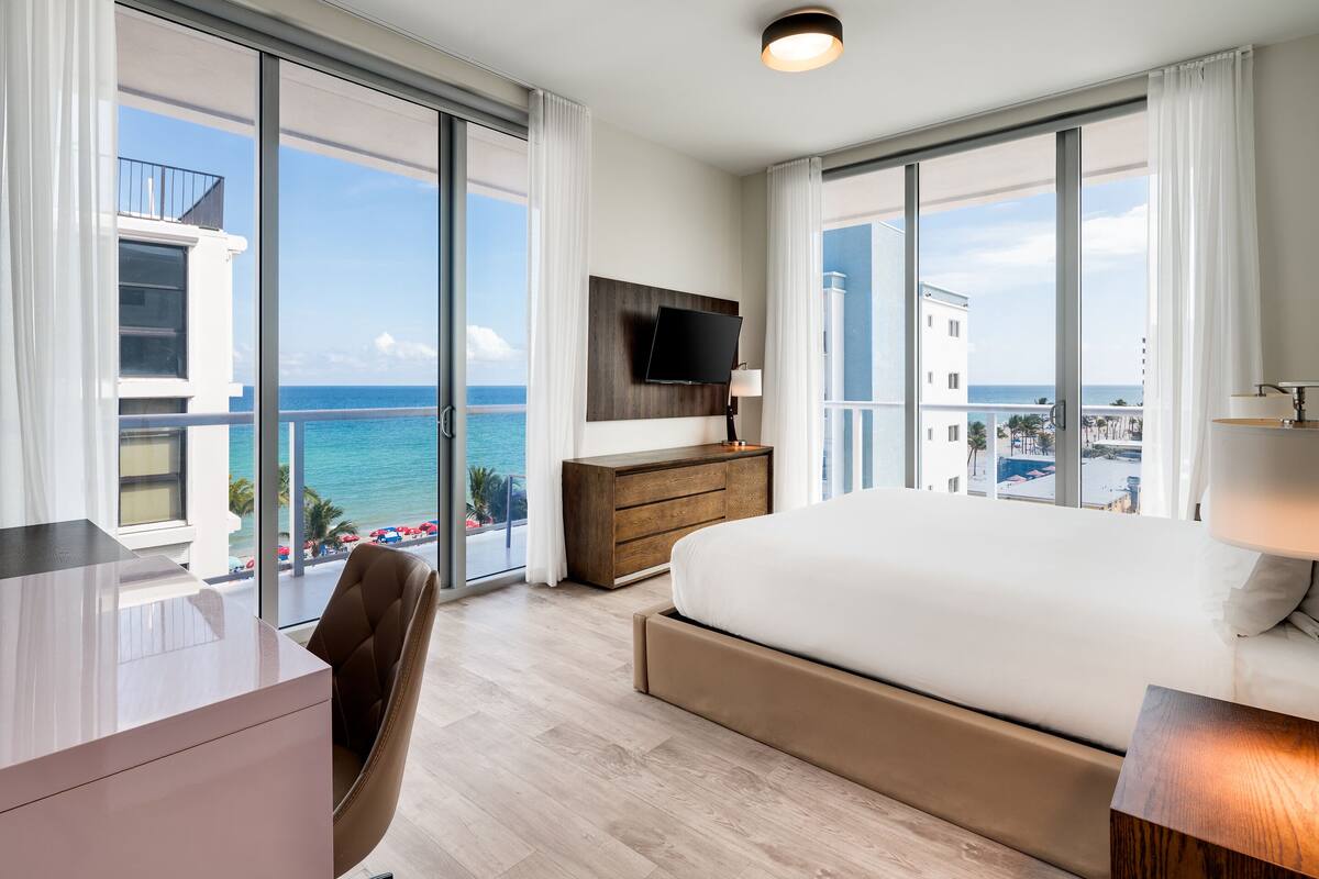 Une chambre d’hôtel moderne, dotée d’un grand lit, d’un bureau et d’une télévision. La chambre dispose de grandes fenêtres offrant une vue sur la mer et les bâtiments environnants.