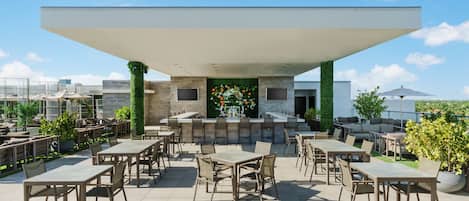 Un área de patio al aire libre con varias mesas y sillas, una barra con fondo verde y vistas de la ciudad.