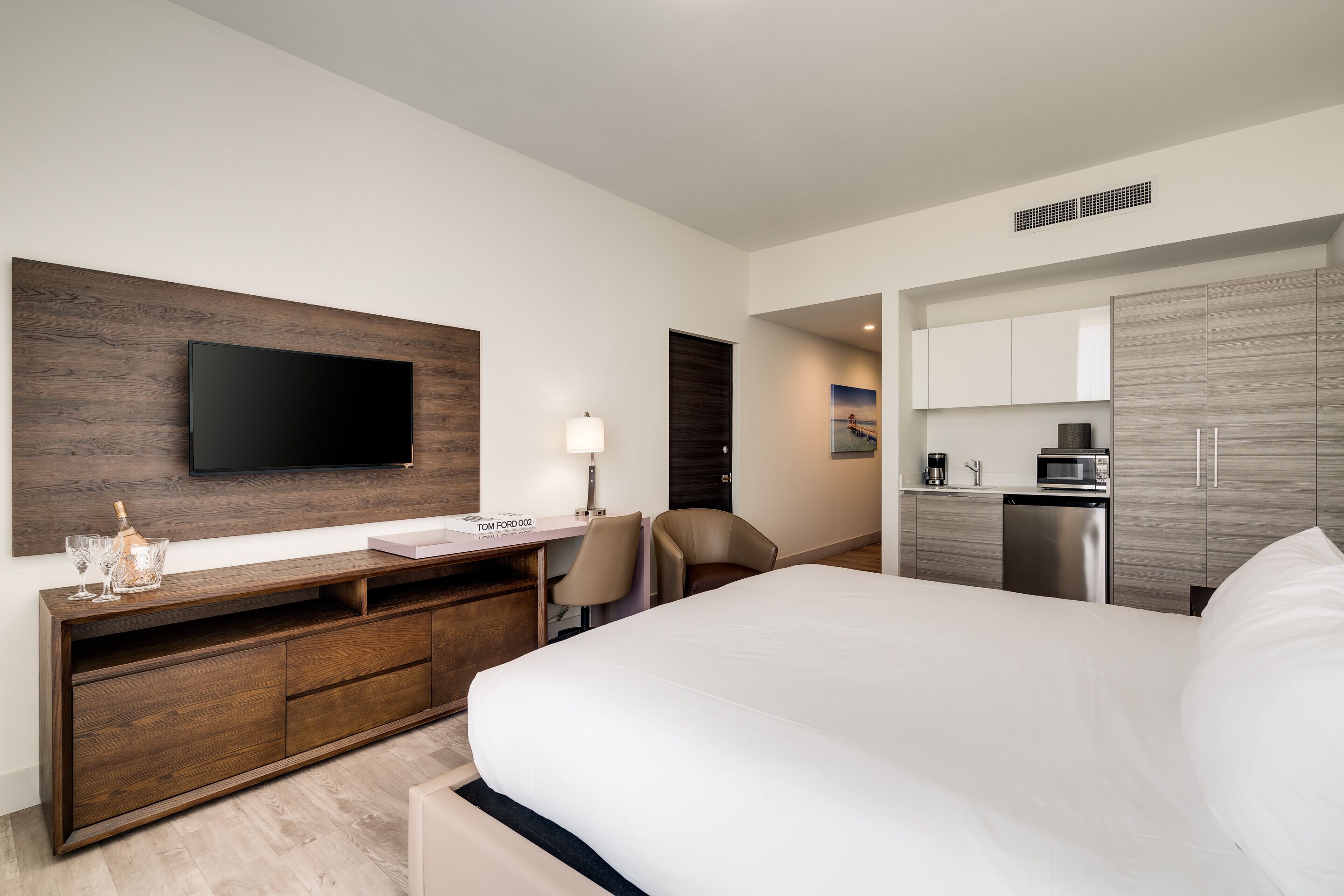 A modern hotel room with a large bed, a flat-screen TV mounted on a wooden panel, a desk with a lamp, a chair, a minibar, and a built-in microwave.