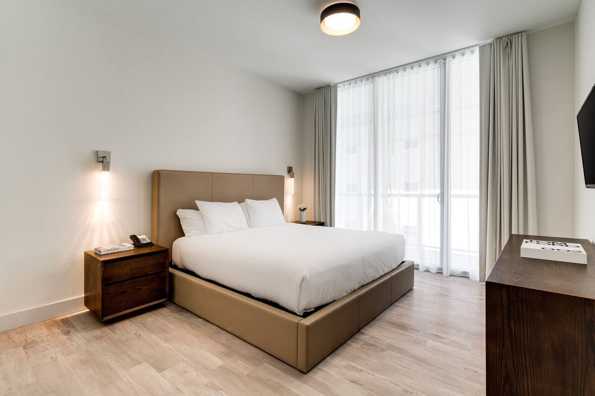 Une chambre d’hôtel moderne avec un grand lit, une table de chevet, un bureau et une grande fenêtre avec des rideaux.
