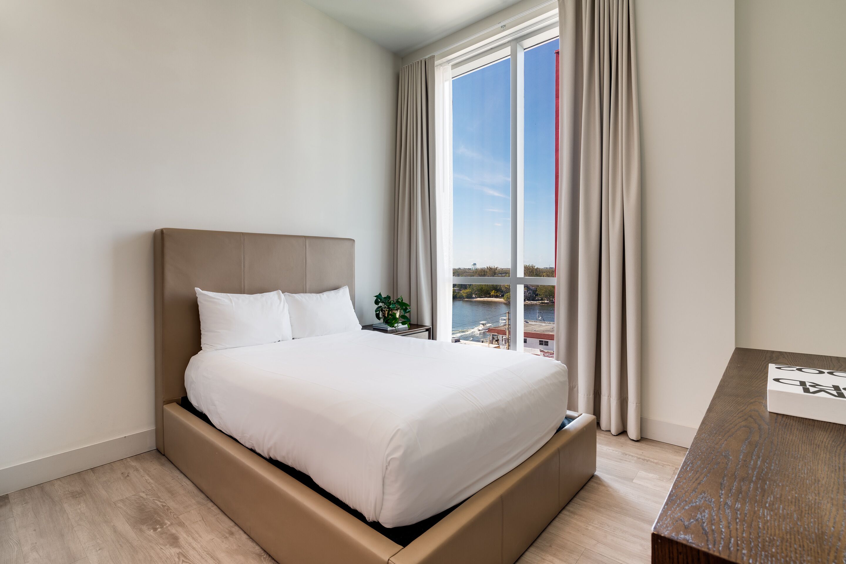 A modern hotel room with a large bed, a wooden nightstand, a view of a body of water, and a cityscape.