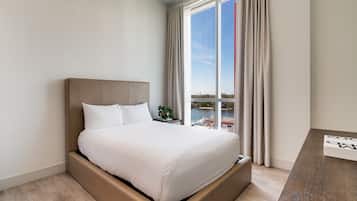 A modern hotel room with a large bed, a wooden nightstand, a view of a body of water, and a cityscape.