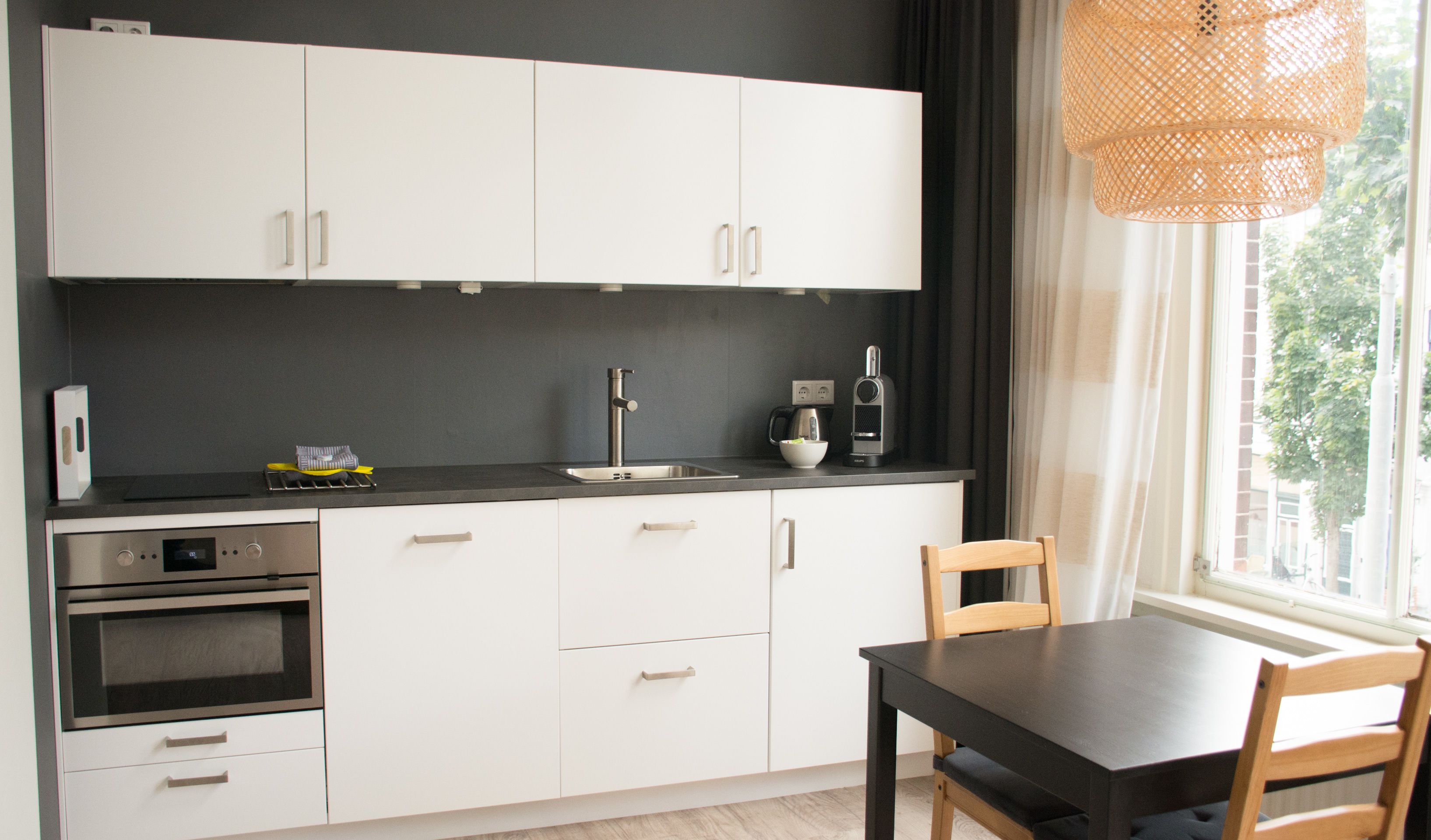 Een moderne keuken met witte kastjes, een zwart aanrecht, een inbouwoven en een eettafel met stoelen.