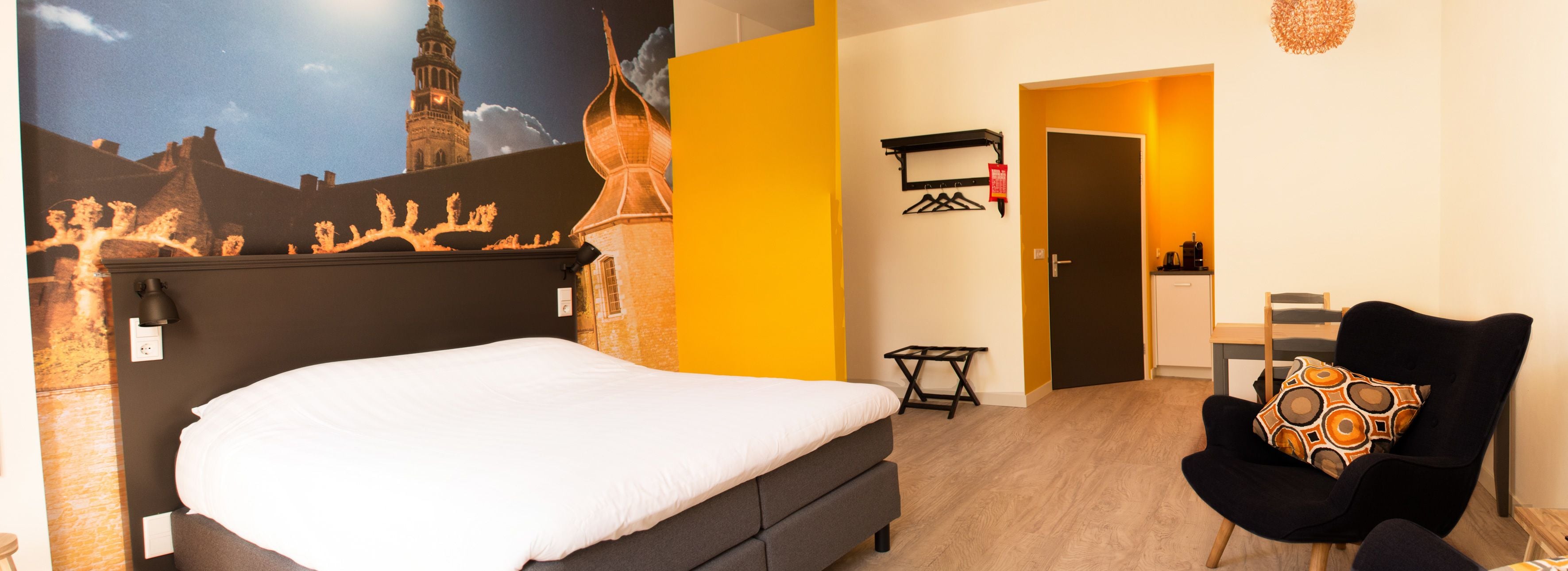 A hotel room with a bed, a chair, a desk, and a wall-mounted shelf.