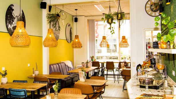 Een restaurantinterieur met meerdere eettafels en stoelen, hangplanten en een toonbank met eten.