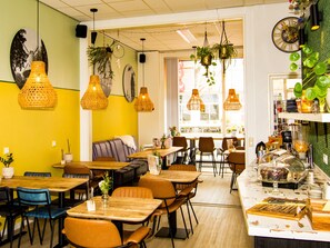 A restaurant interior with multiple dining tables and chairs, hanging plants, and a counter with food items. - ApartHotel Waepen van Middelburg (Middelburg)