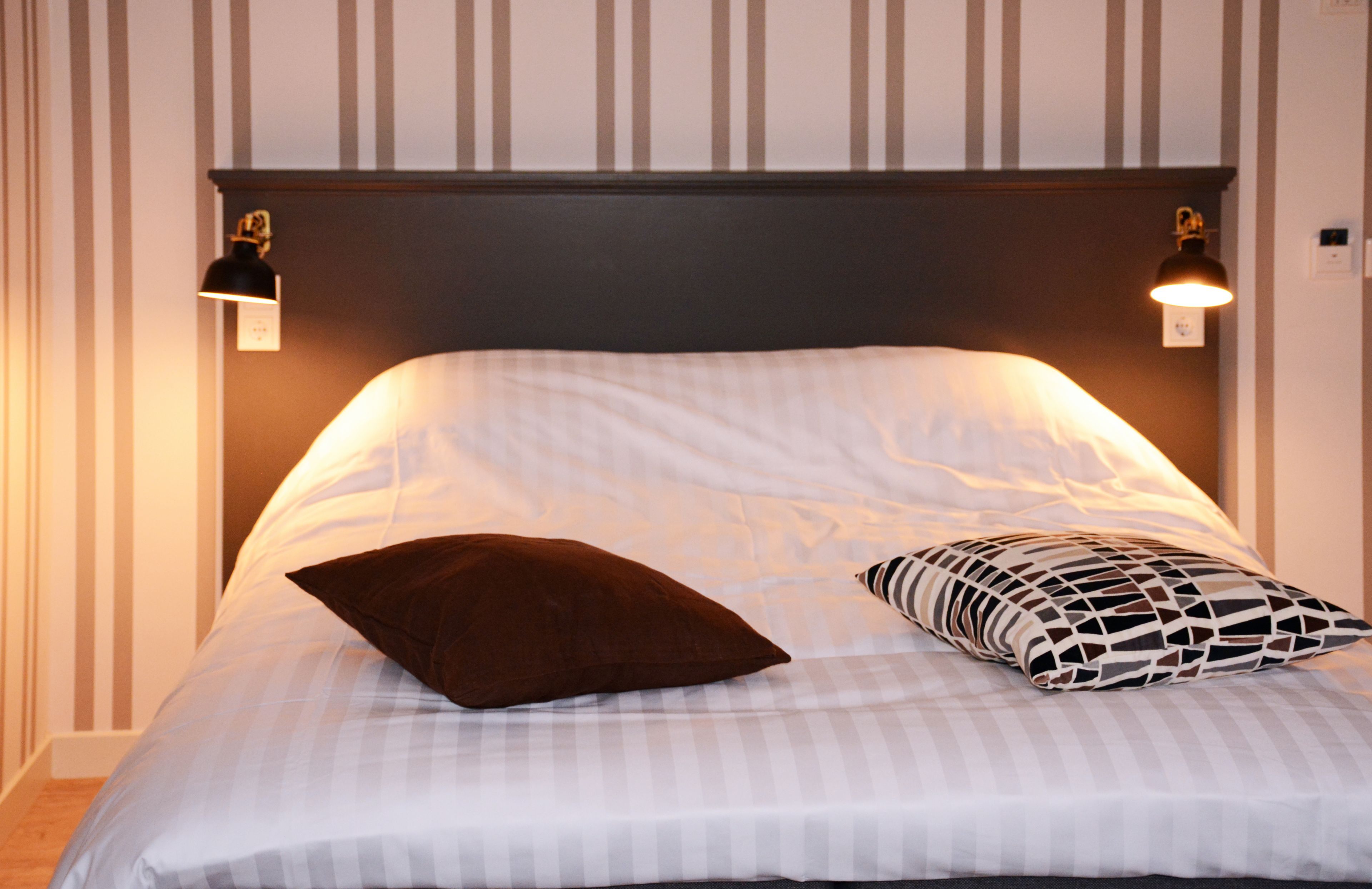 A neatly made bed with striped bedding, two wall-mounted reading lamps, and a dark brown pillow.