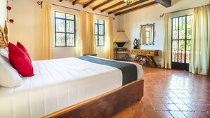 Ein geräumiges Schlafzimmer mit einem großen Bett, einem hölzernen Kopfteil und einem rustikalen Fliesenboden. Zwei Fenster mit Vorhängen, ein Spiegel und ein kleiner Tisch mit einer Lampe sind ebenfalls vorhanden.