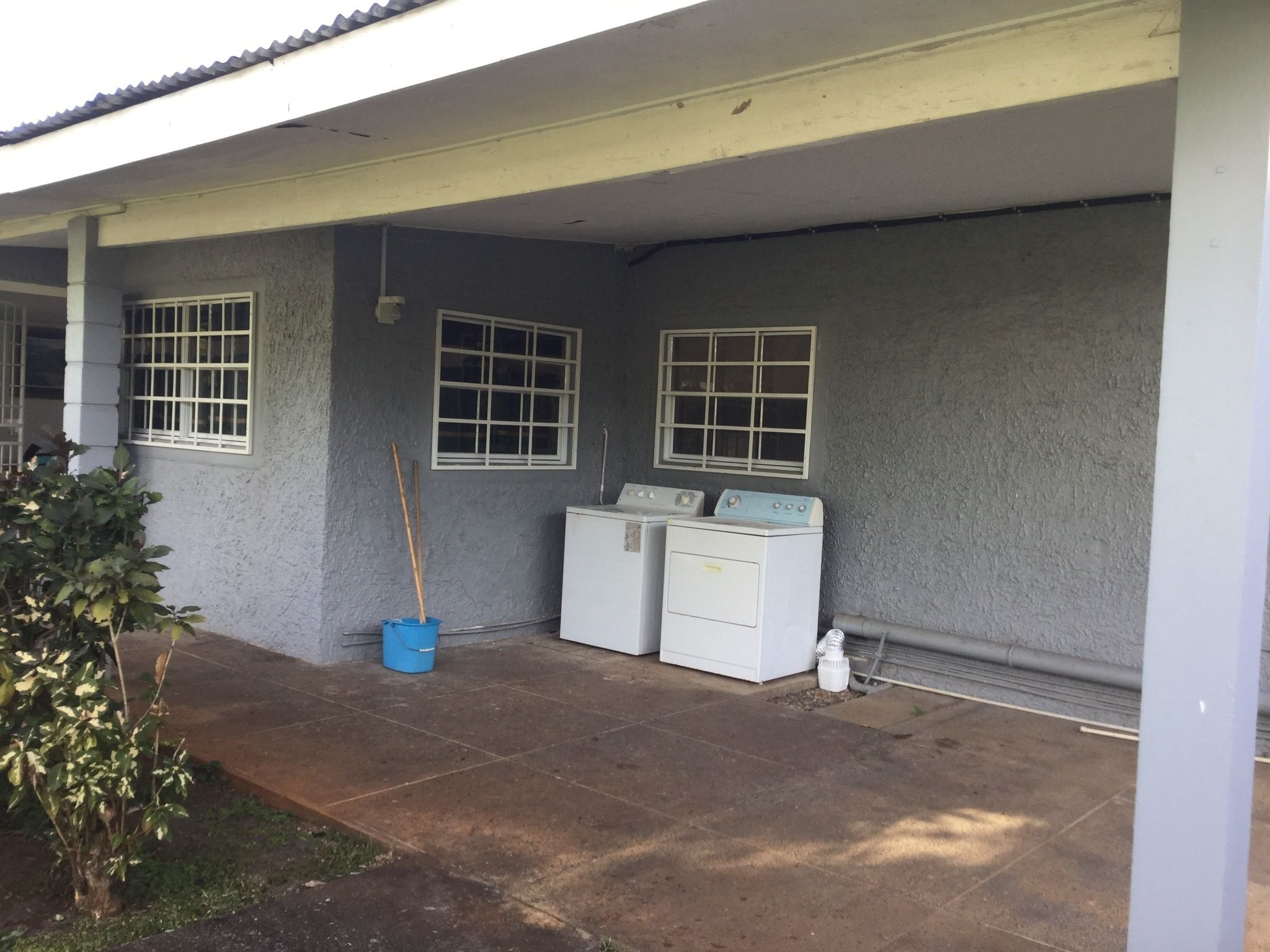 A laundry area with two washing machines, a blue bucket, and a broom leaning against the wall.