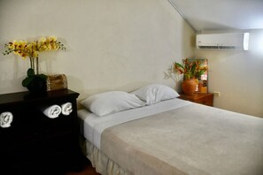 A neatly made bed with white linens, a small bedside table with a potted plant, and a wall-mounted air conditioner. - Ella's Park View Townhouse Crown Point (Crown Point)