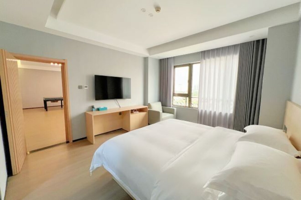 A modern hotel room with a large bed, a desk with a flat-screen TV, a sofa, and a window with curtains.