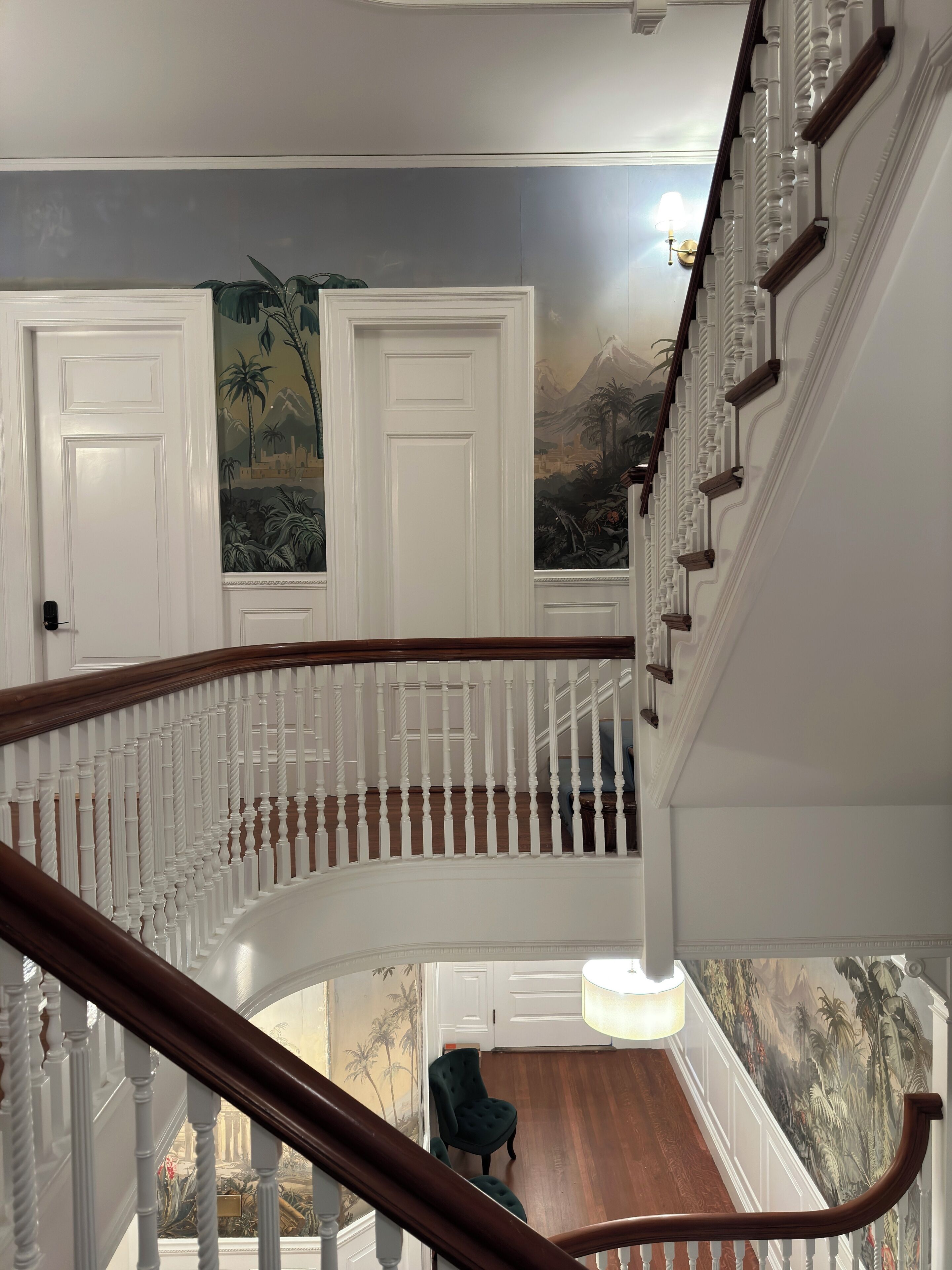 Een statige trap met witte balustrades en een houten leuning, die uitkomt op een plateau met een met groen bekleden stoel en een muurschildering van tropische landschappen.