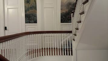 Een statige trap met witte balustrades en een houten leuning, die uitkomt op een plateau met een met groen bekleden stoel en een muurschildering van tropische landschappen.