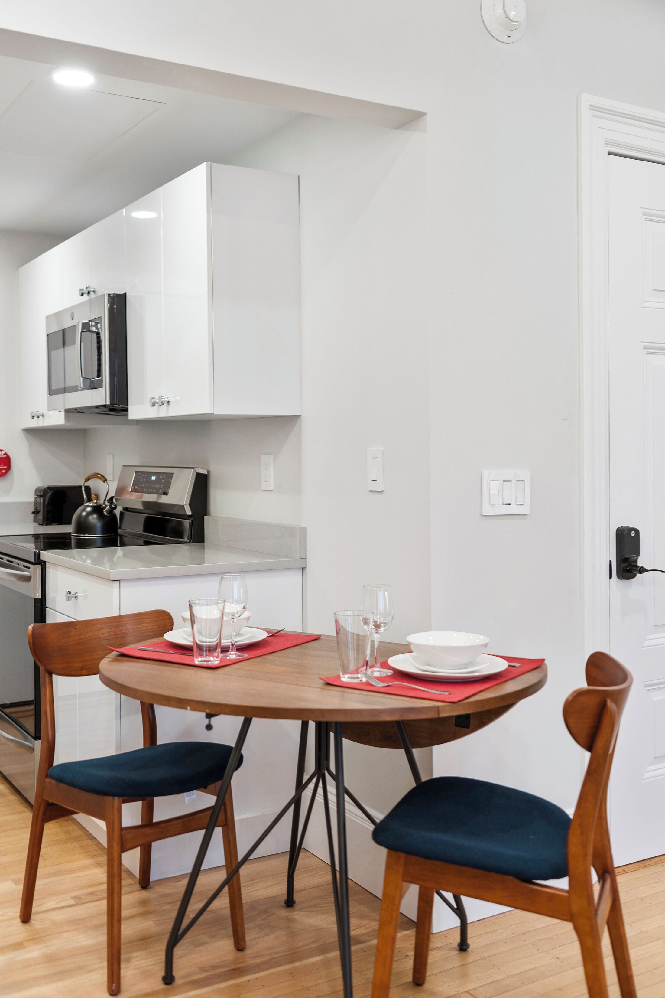 Cocina compacta con muebles blancos, un microondas y un hervidor en la encimera. Hay una mesa de comedor con dos sillas, cada una con un cojín oscuro.