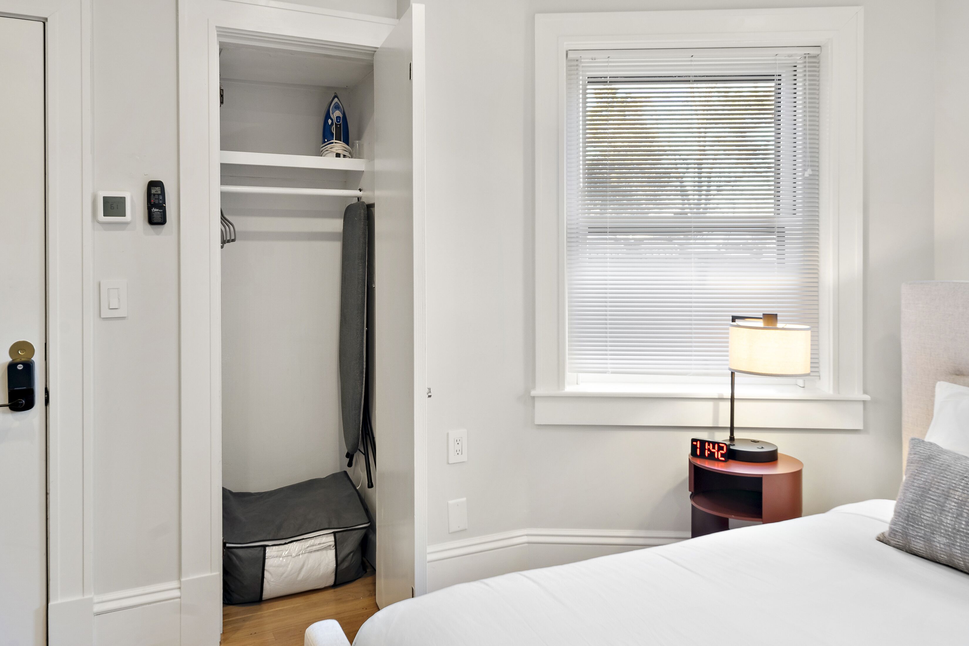 A bedroom with a bed, a nightstand with a lamp, a window with blinds, a closet with shelves, and a small seating area.
