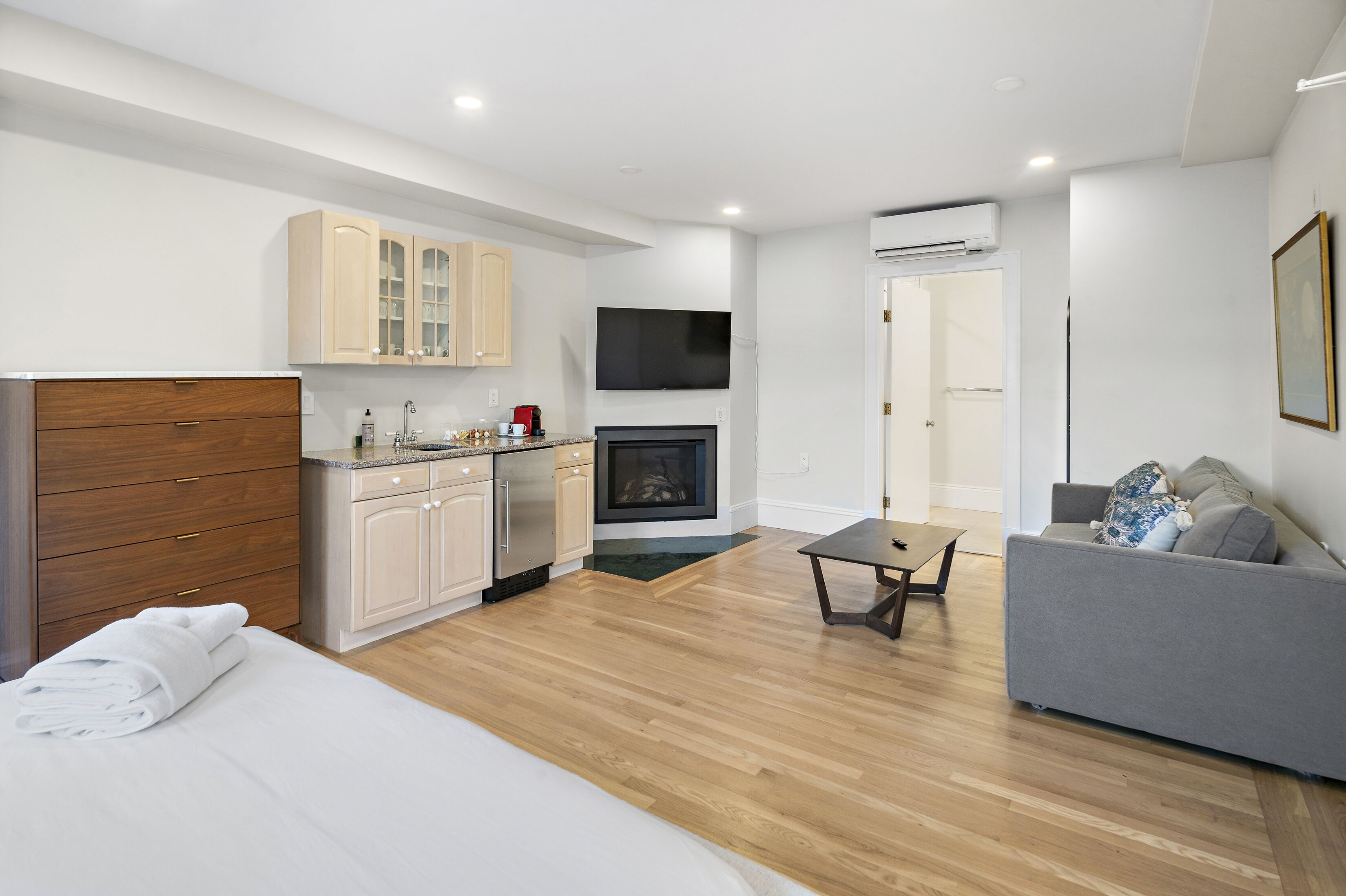 Ein modernes Hotelzimmer mit Holzboden, einem Bett mit weißen Bettwäsche, einem grauen Sofa, einem kleinen Tisch, einer Küchenzeile mit Schränken und einem Flachbildfernseher sowie einer Klimaanlage an der Wand.