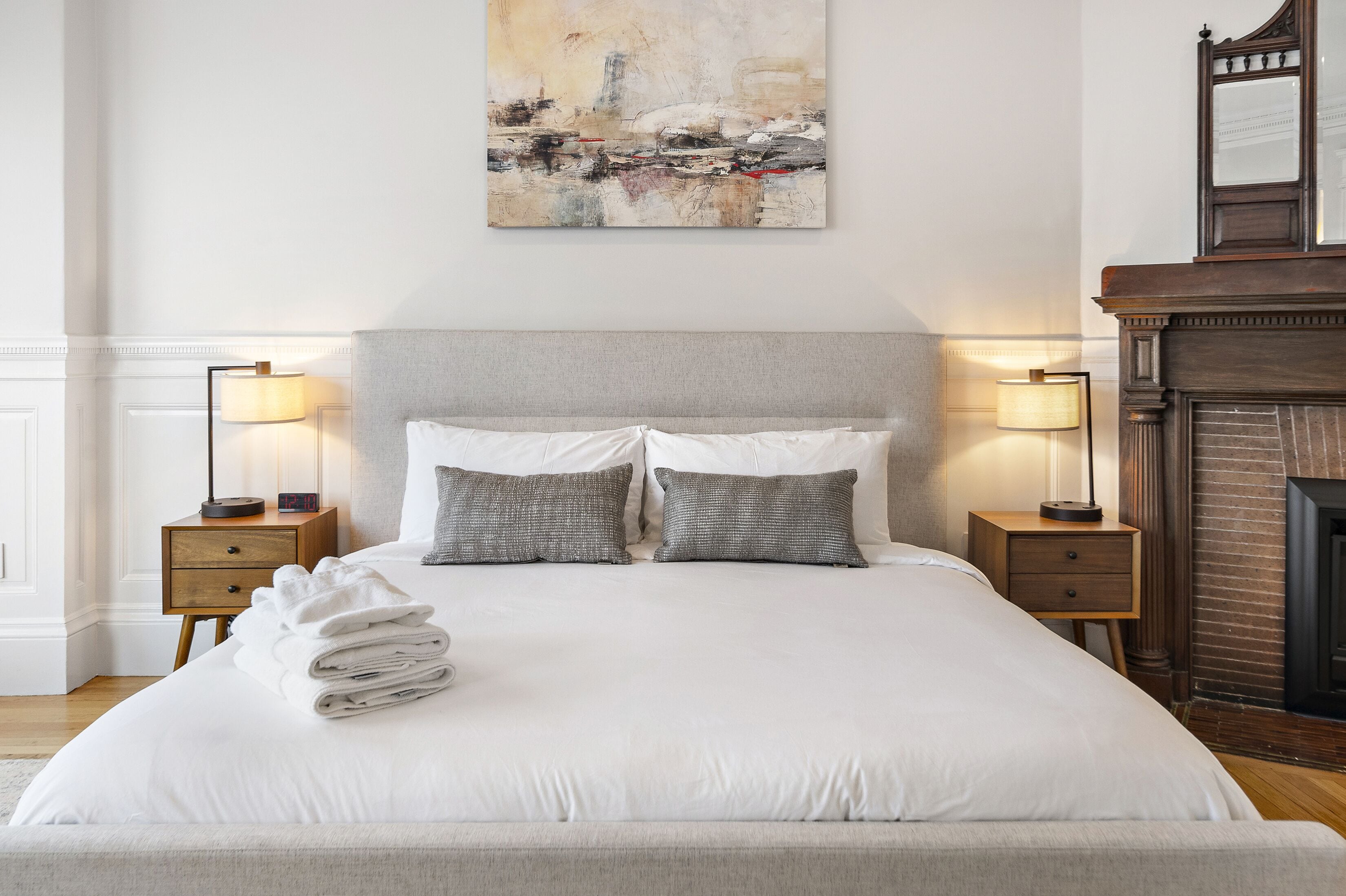 Una cama bien tendida con sábanas blancas y dos almohadas, flanqueada por mesitas de noche con lámparas, en una habitación con chimenea y un cuadro de paisaje urbano en la pared.