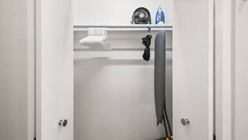 A room with a white door, a wooden floor, a towel hanging on a rack, a fan, and an ironing board.