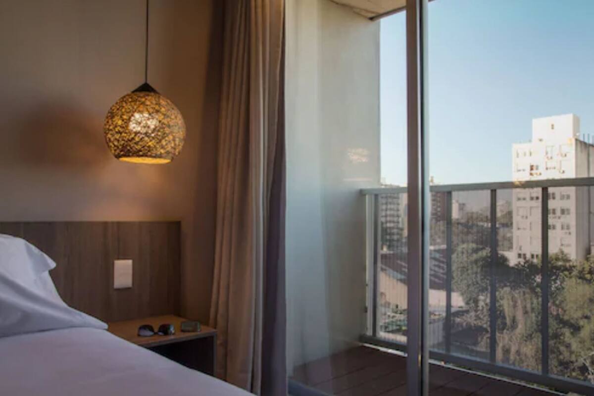 A hotel room with a bed, a bedside table with glasses, a hanging lamp, and a view of buildings through large windows.