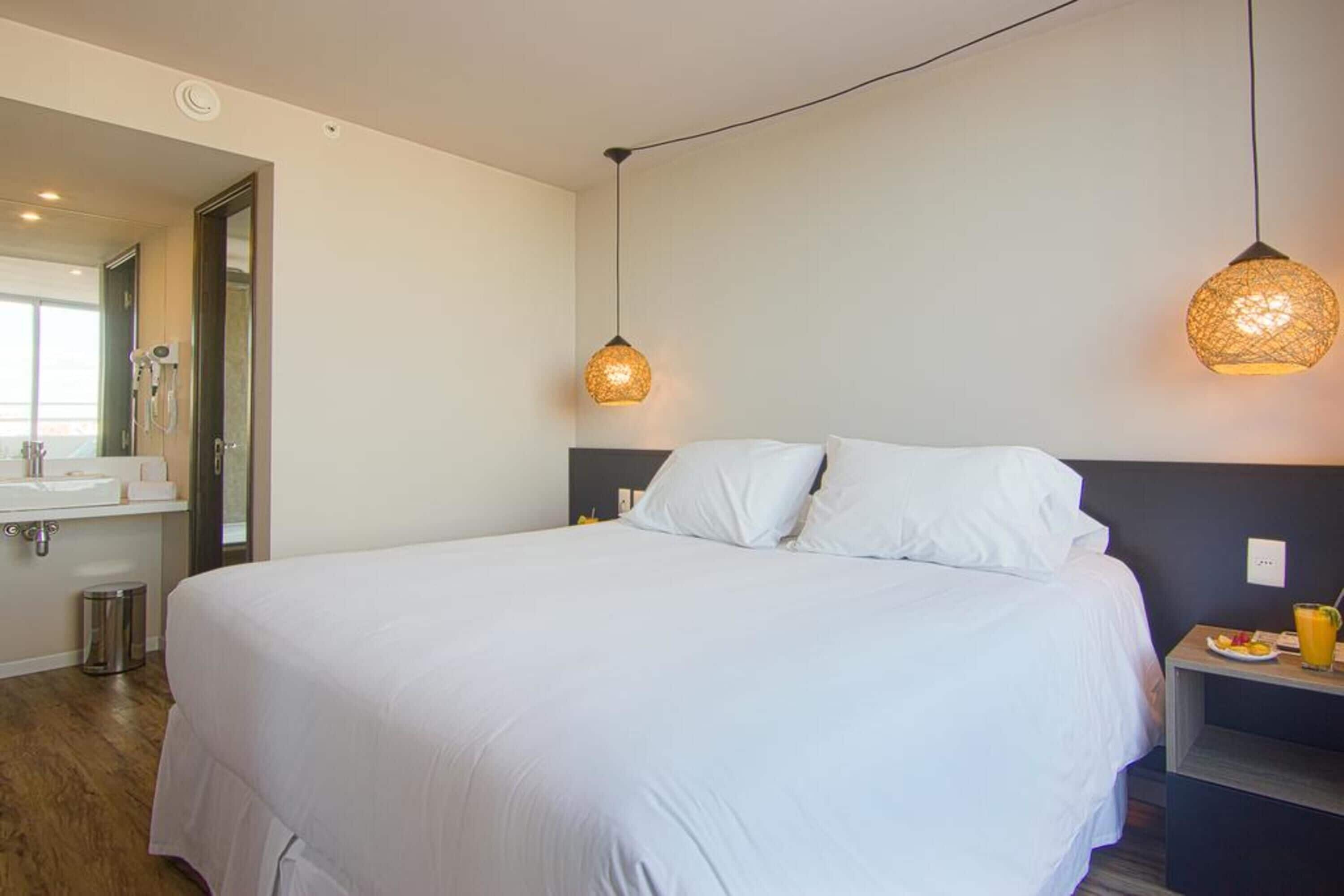 Una habitación de hotel con una cama grande, una mesita de noche con un vaso de jugo de naranja y un baño con lavamanos y espejo.