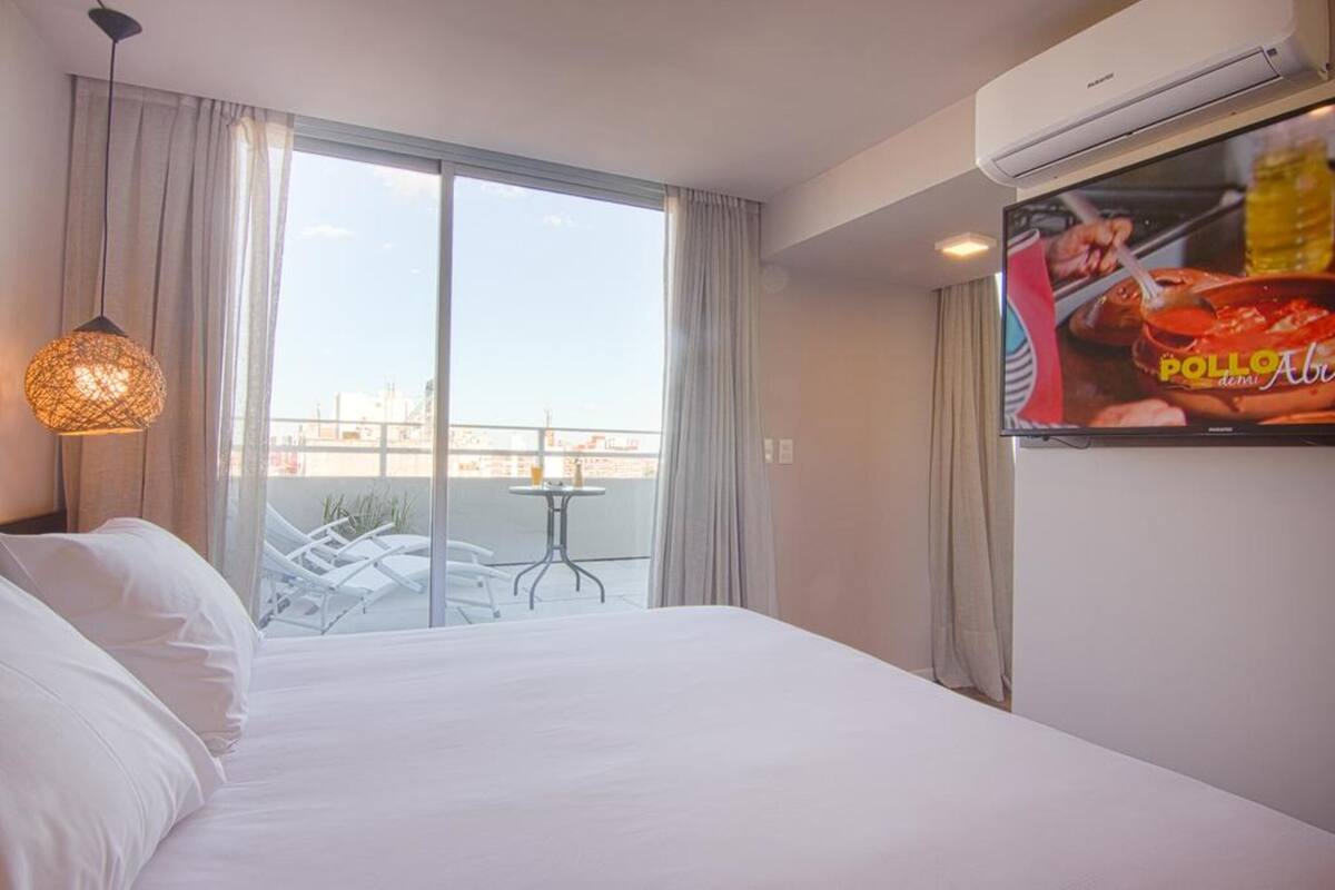 A hotel room with a bed, a TV, a balcony with a table and chairs, and a view of the city.