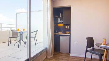 Cocina compacta con refrigerador, microondas y cafetera. Un área de comedor con una mesa y sillas está junto a un ventanal amplio.