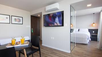 A modern hotel room with a bed, a TV, a dining area with a table and chairs, and a breakfast tray with orange juice.