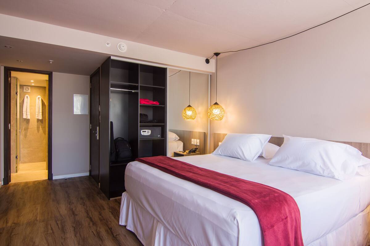 A hotel room with a large bed, a wardrobe, a bedside table with a lamp, and a door leading to a bathroom.
