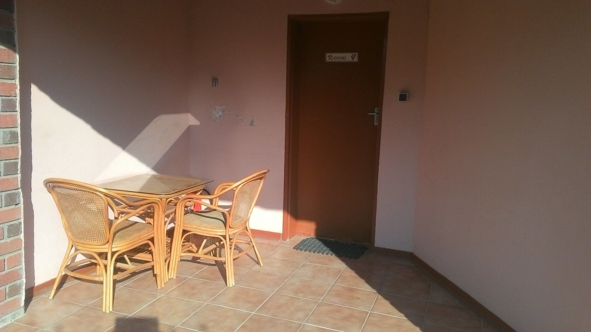A small outdoor seating area with a table and four wicker chairs, a door labeled "Room 9", and a tiled floor.