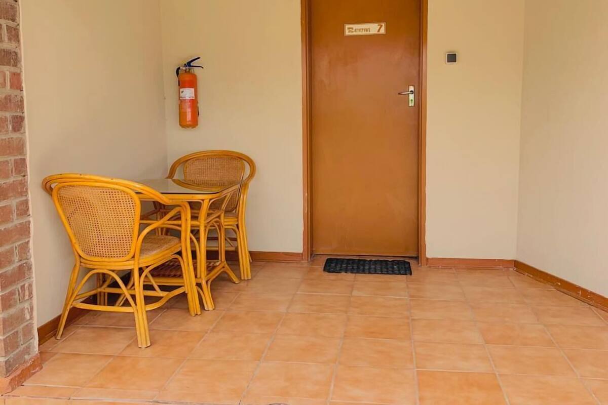 A room with a wooden door labeled "Room 7," a small table with two wicker chairs, a fire extinguisher on the wall, and a tiled floor.
