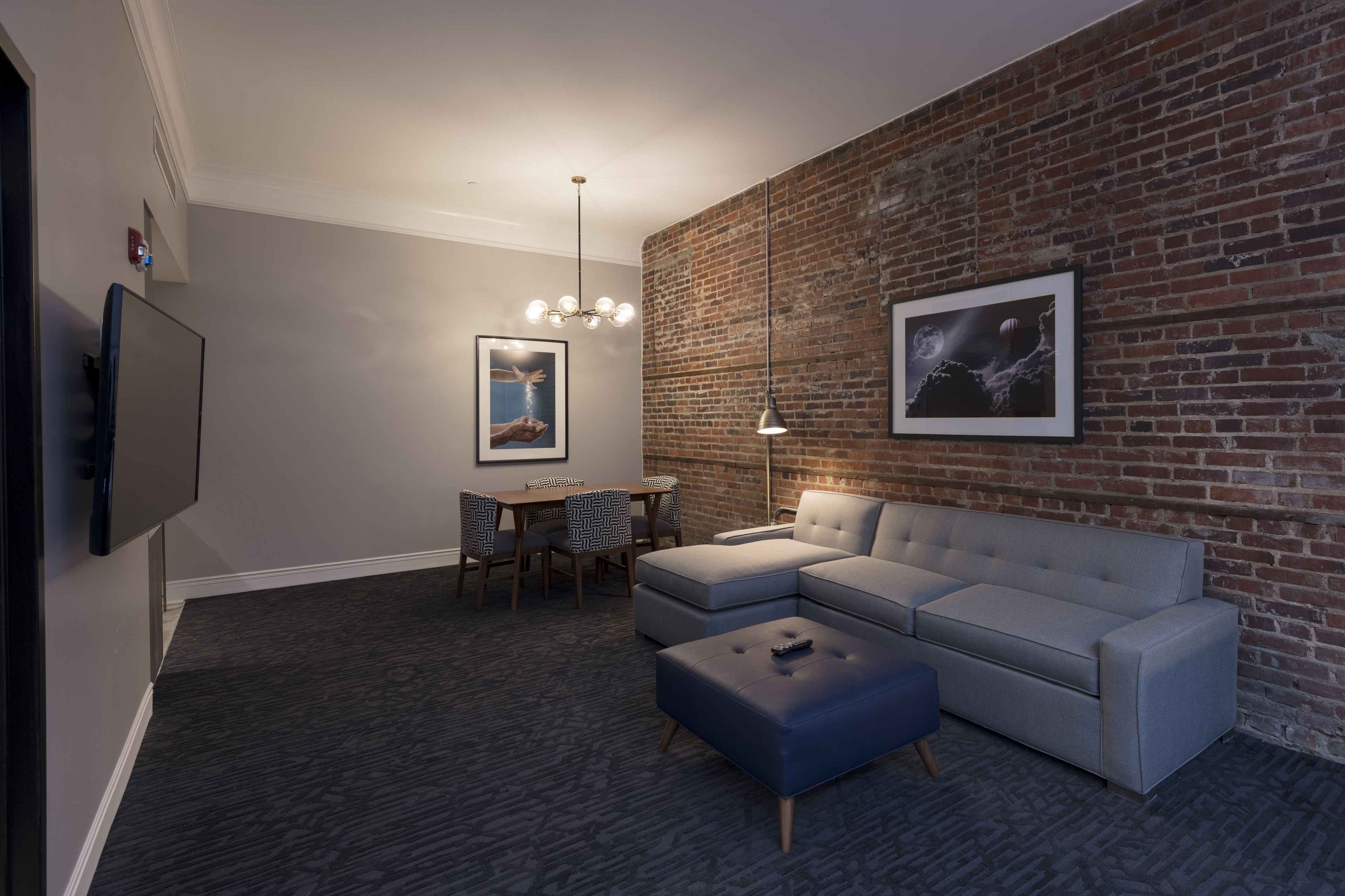 Una sala moderna con un sofá seccional gris, una mesita con sillas, un televisor montado en la pared y una pared de ladrillo con obras de arte enmarcadas.