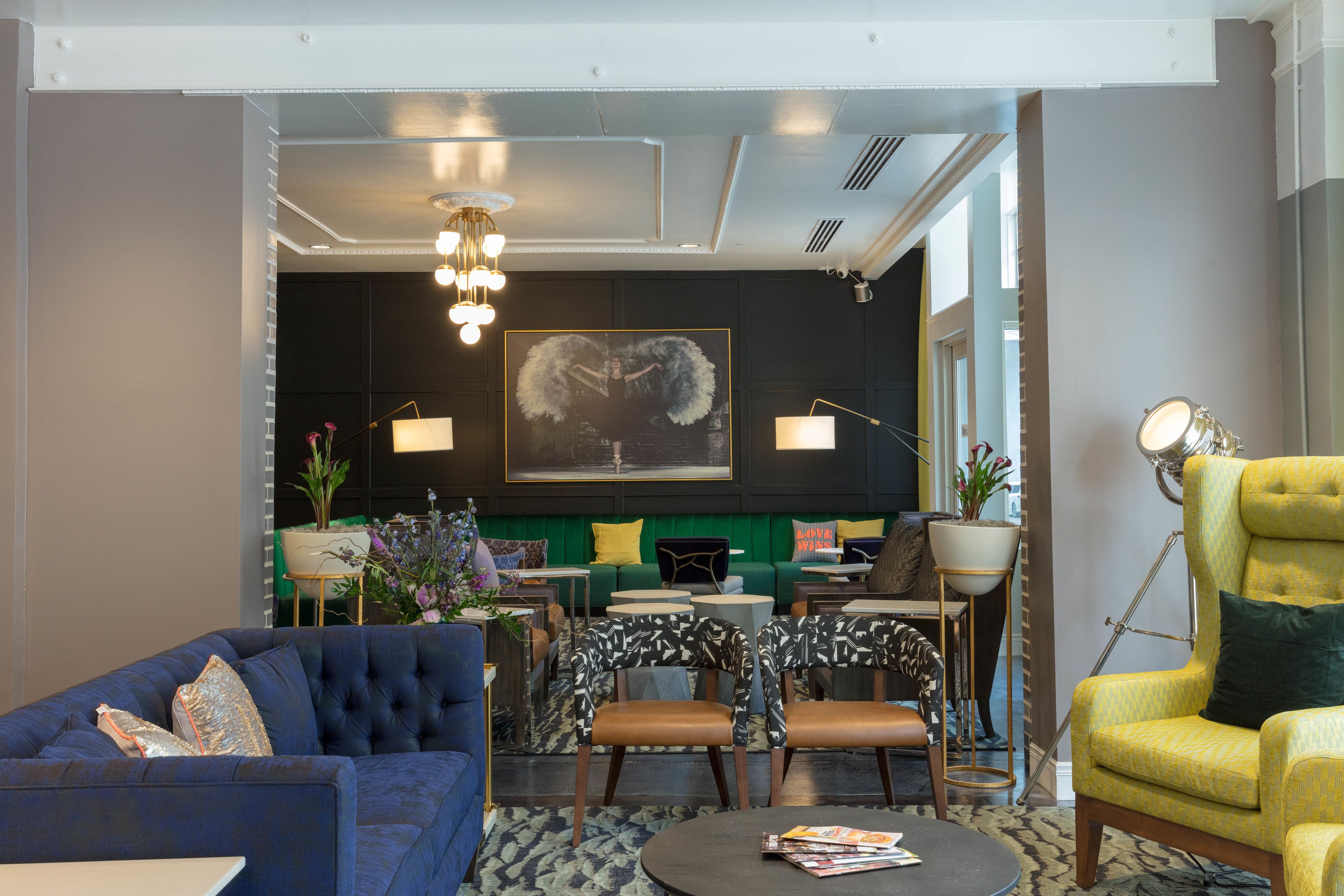 Un área de estar moderna con un sofá azul, un sillón amarillo y un juego de sofá verde. Hay una mesa de centro redonda con revistas, una lámpara de pie y un cuadro en la pared.