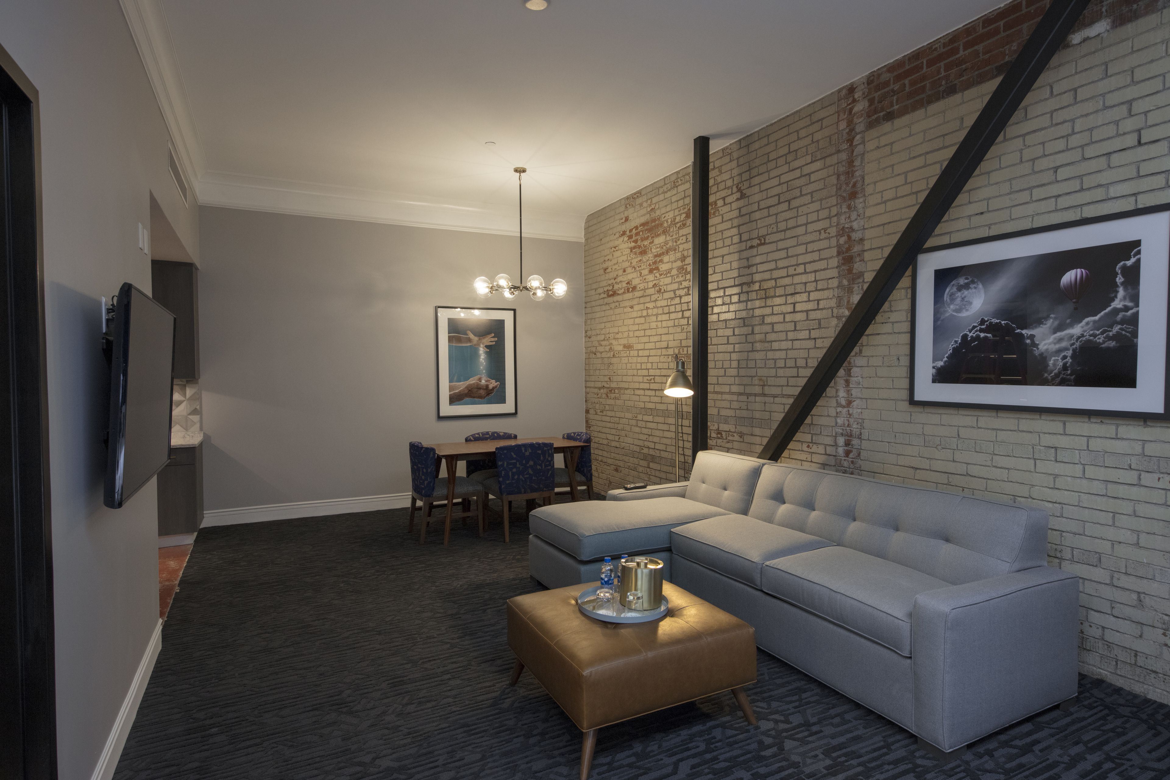A modern living room with a grey sofa, a coffee table, a dining area with a table and chairs, and a brick wall with a framed picture.