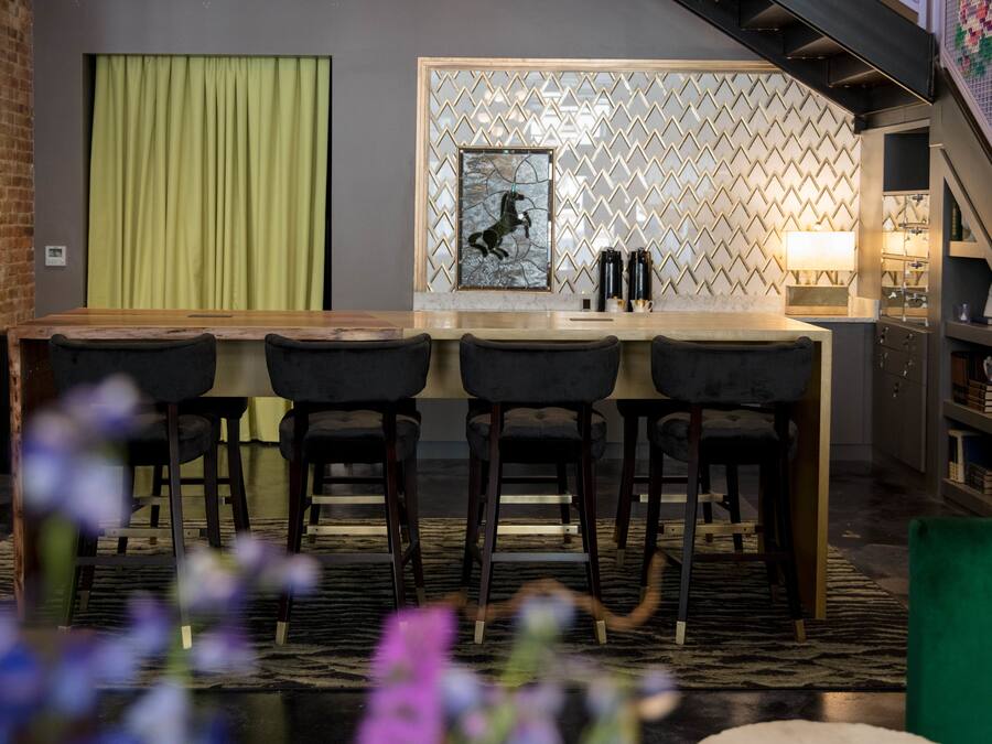 A bar area with a wooden counter, three bar stools, a green curtain, a patterned wall, and a staircase.
