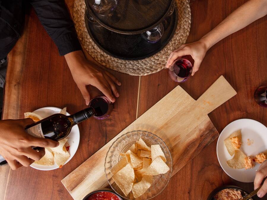 A wooden table with a bottle of wine, glasses, tortilla chips, and salsa.